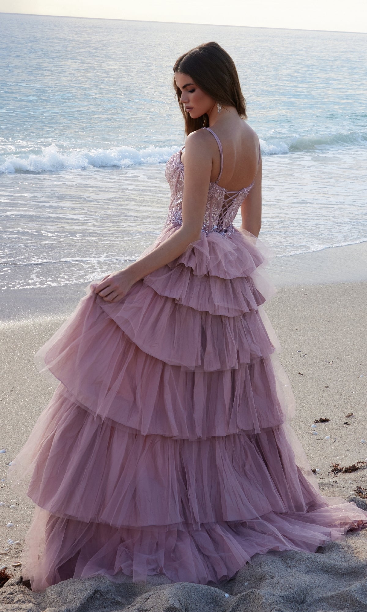 A woman in the Nox Anabel Y1544 Overskirt Long Lace Prom Dress stands on a sandy beach, facing the ocean as gentle waves roll in.
